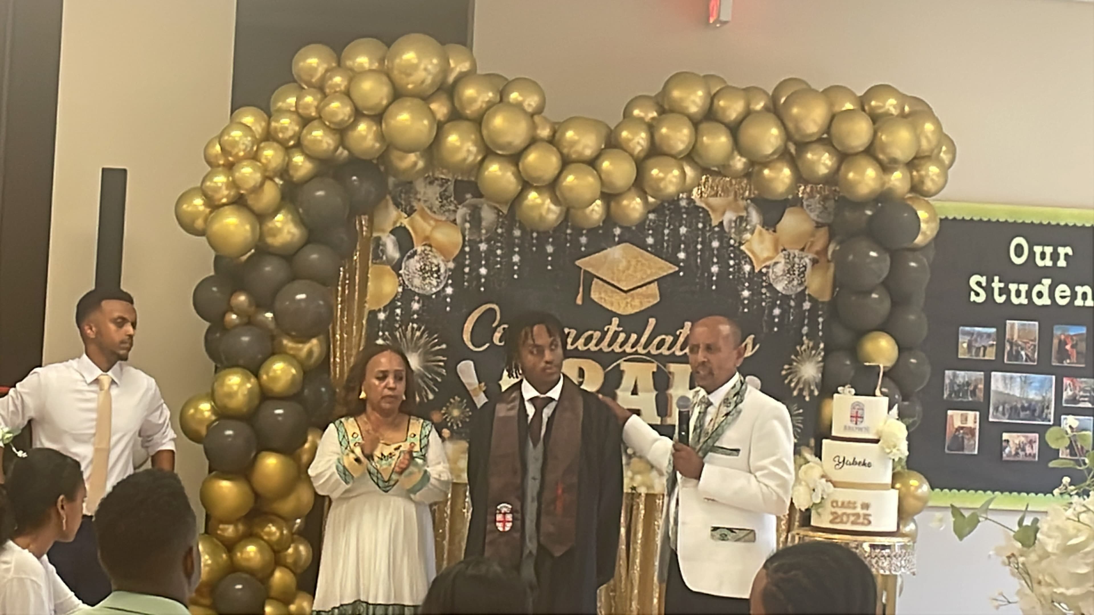 A graduate in a gown stands with family before a gold and black balloon arch.