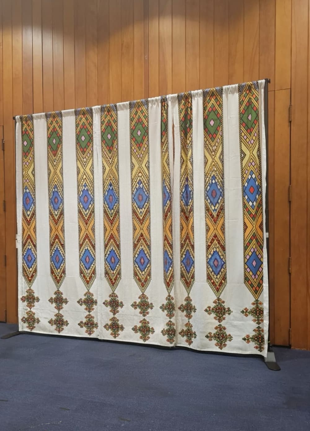White curtain featuring colorful vertical diamond patterns and cross motifs against a wooden wall.