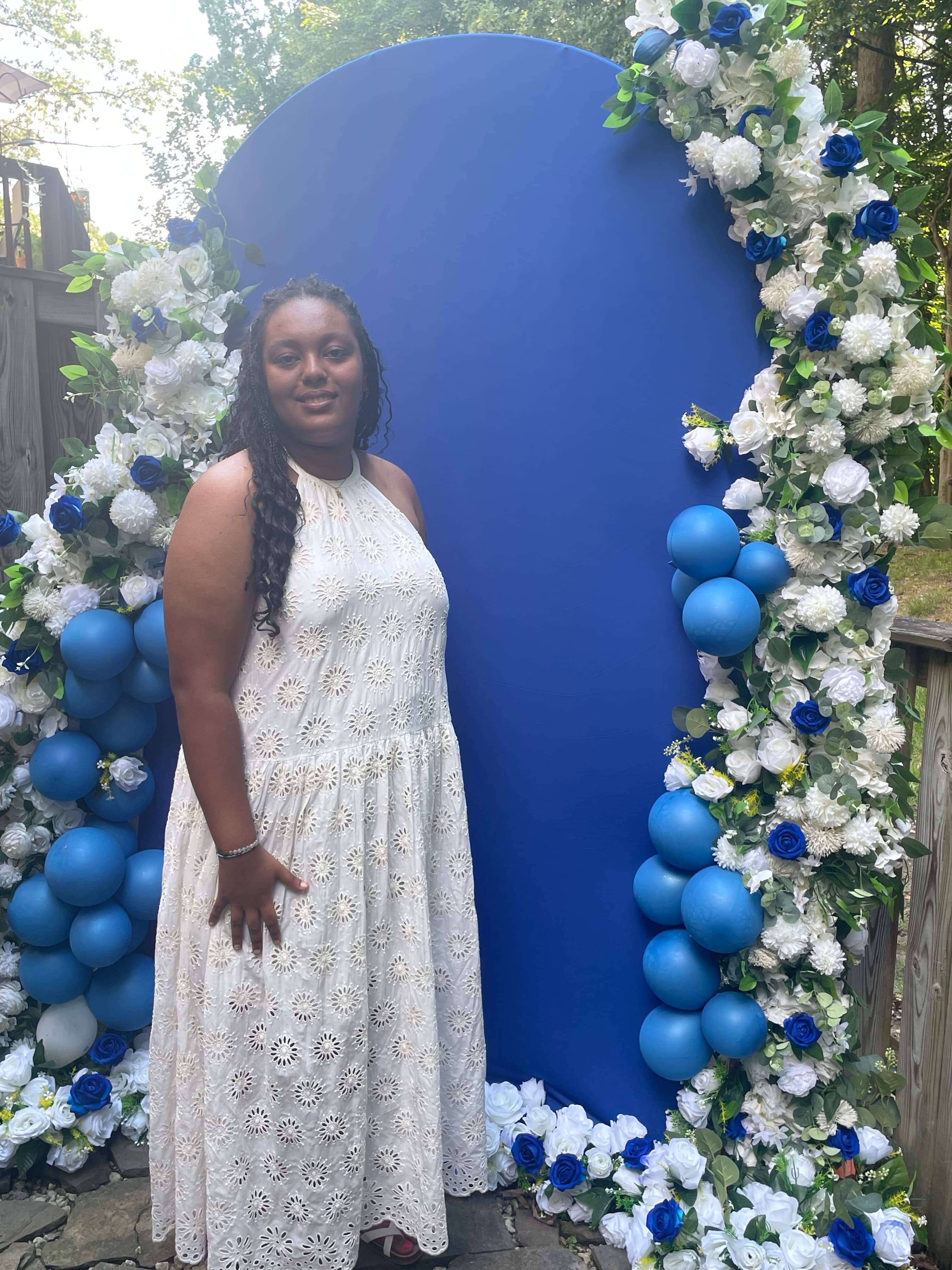 Smiling woman in a white eyelet dress stands before a blue floral and balloon backdrop.