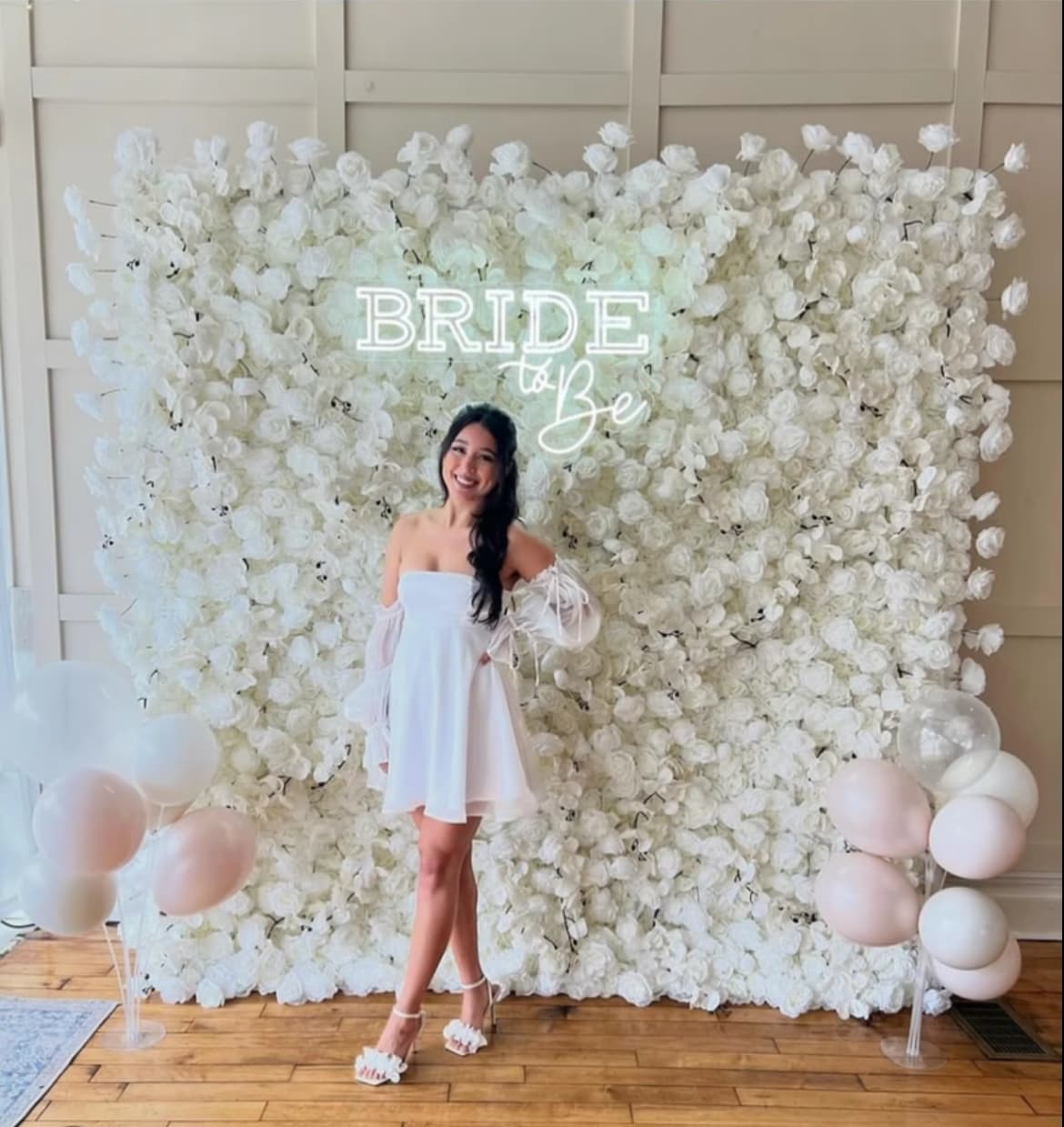 Woman in white dress posing before white flower wall with "Bride to Be" neon sign.