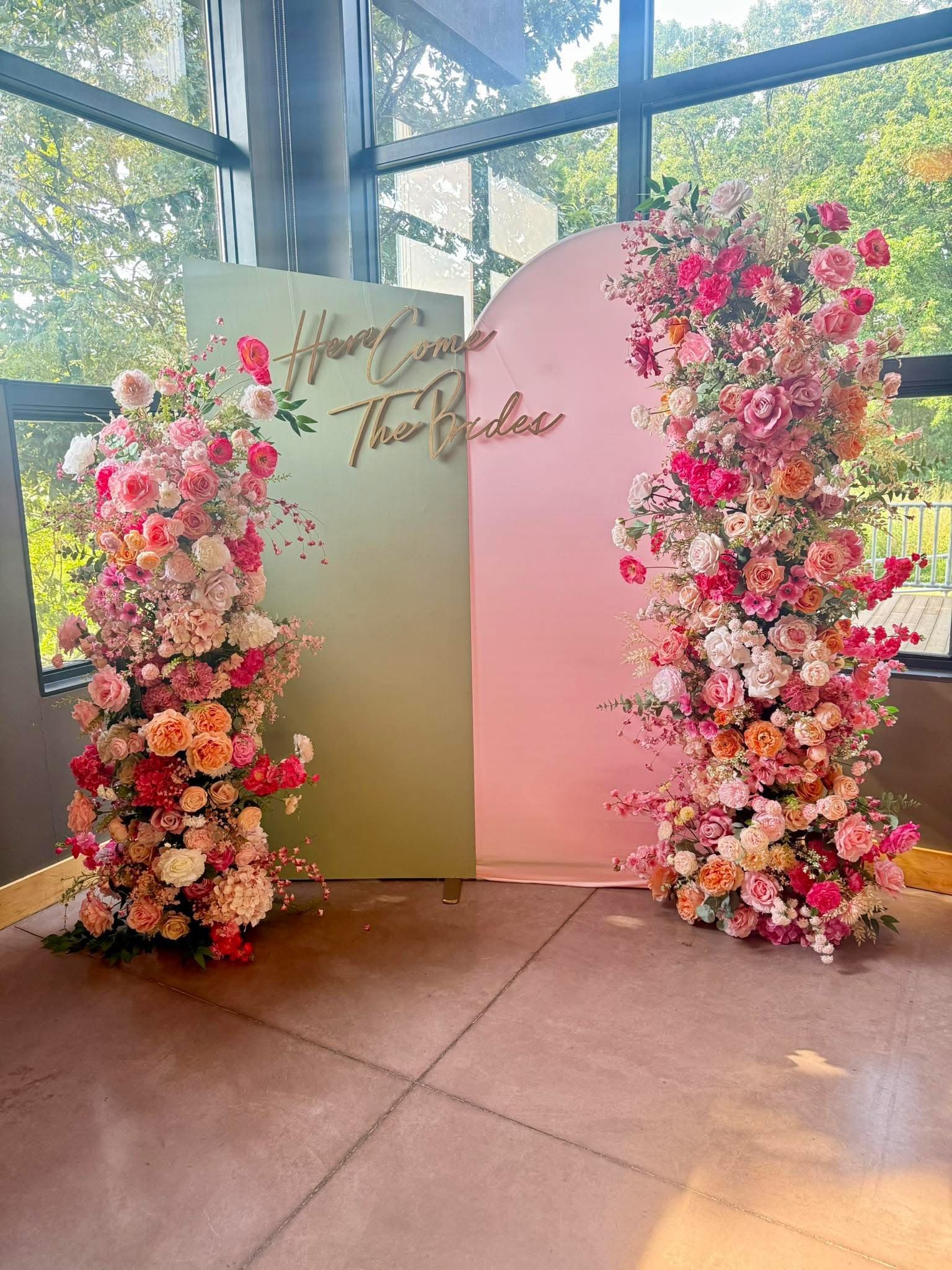 Green and pink backdrop with lush floral pillars and gold "Here Come The Brides" lettering.