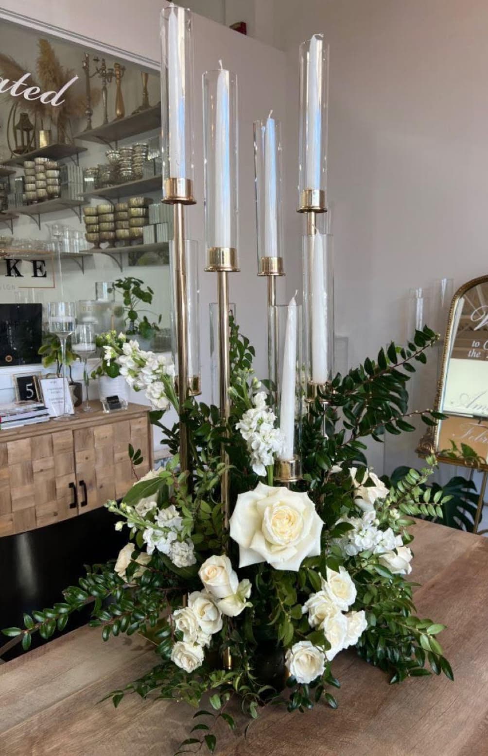 Elegant white rose floral centerpiece with greenery and tall gold candle holders in glass cylinders.