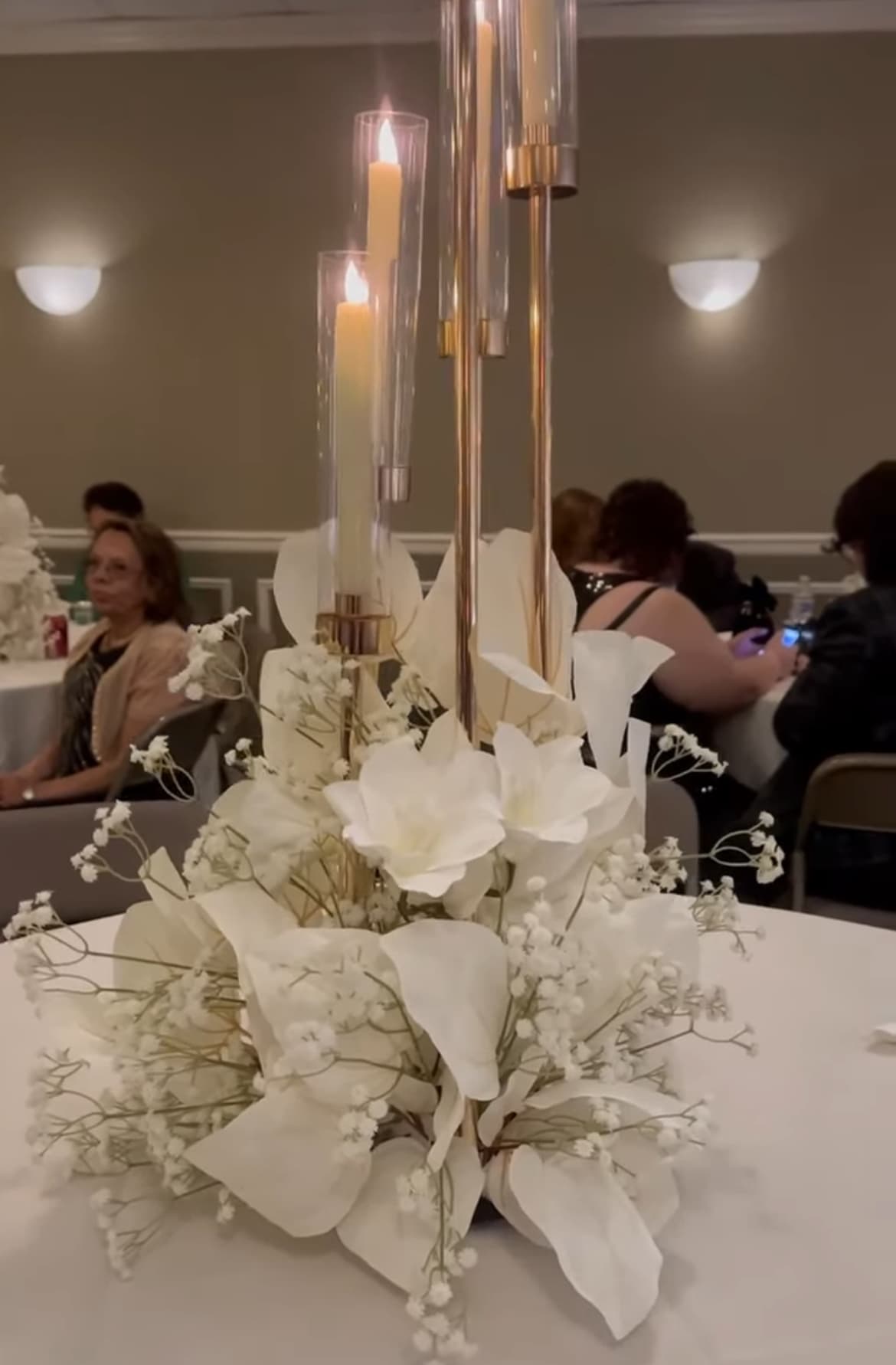 Elegant white floral centerpiece with tall gold candle holders and lit candles on a table.