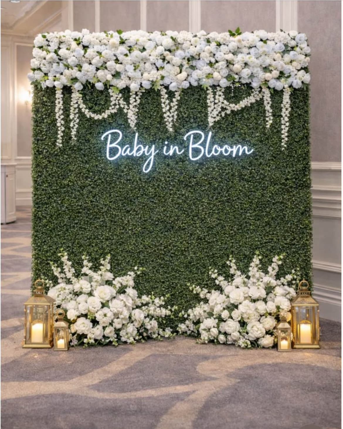 Greenery wall with white flowers, gold lanterns, and a neon "Baby in Bloom" sign.