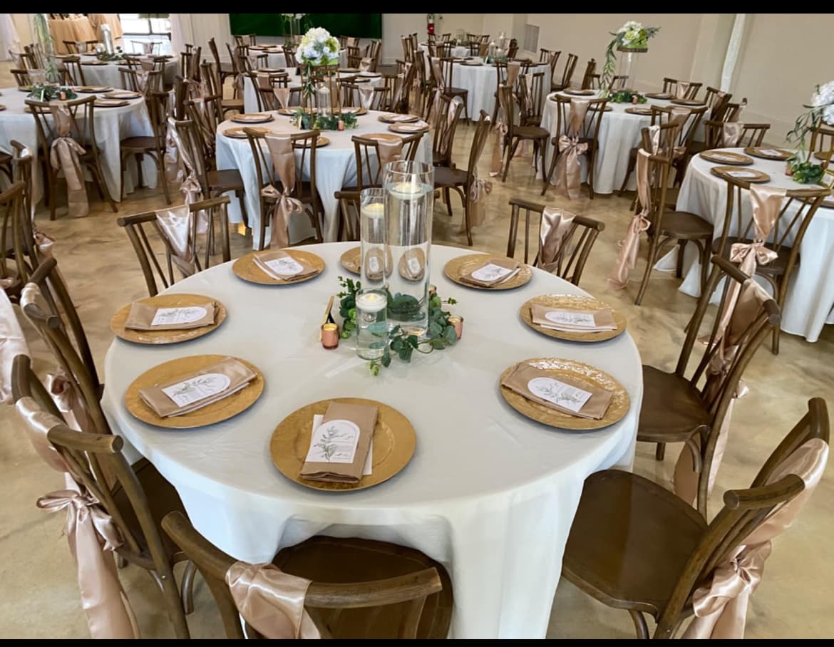 Elegant reception hall with round tables, gold chargers, and wooden chairs featuring satin bows.