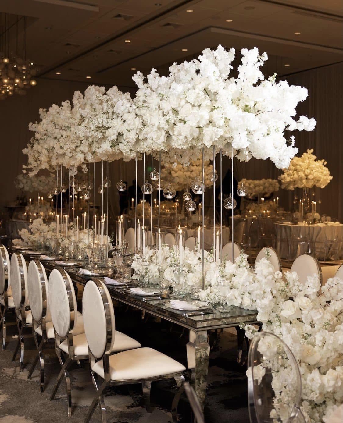 Mirrored banquet table decorated with towering white floral clouds, hanging candles, and white chairs.