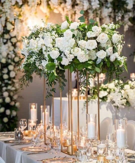 Lush white floral centerpiece on a tall gold stand above a candlelit wedding reception table.