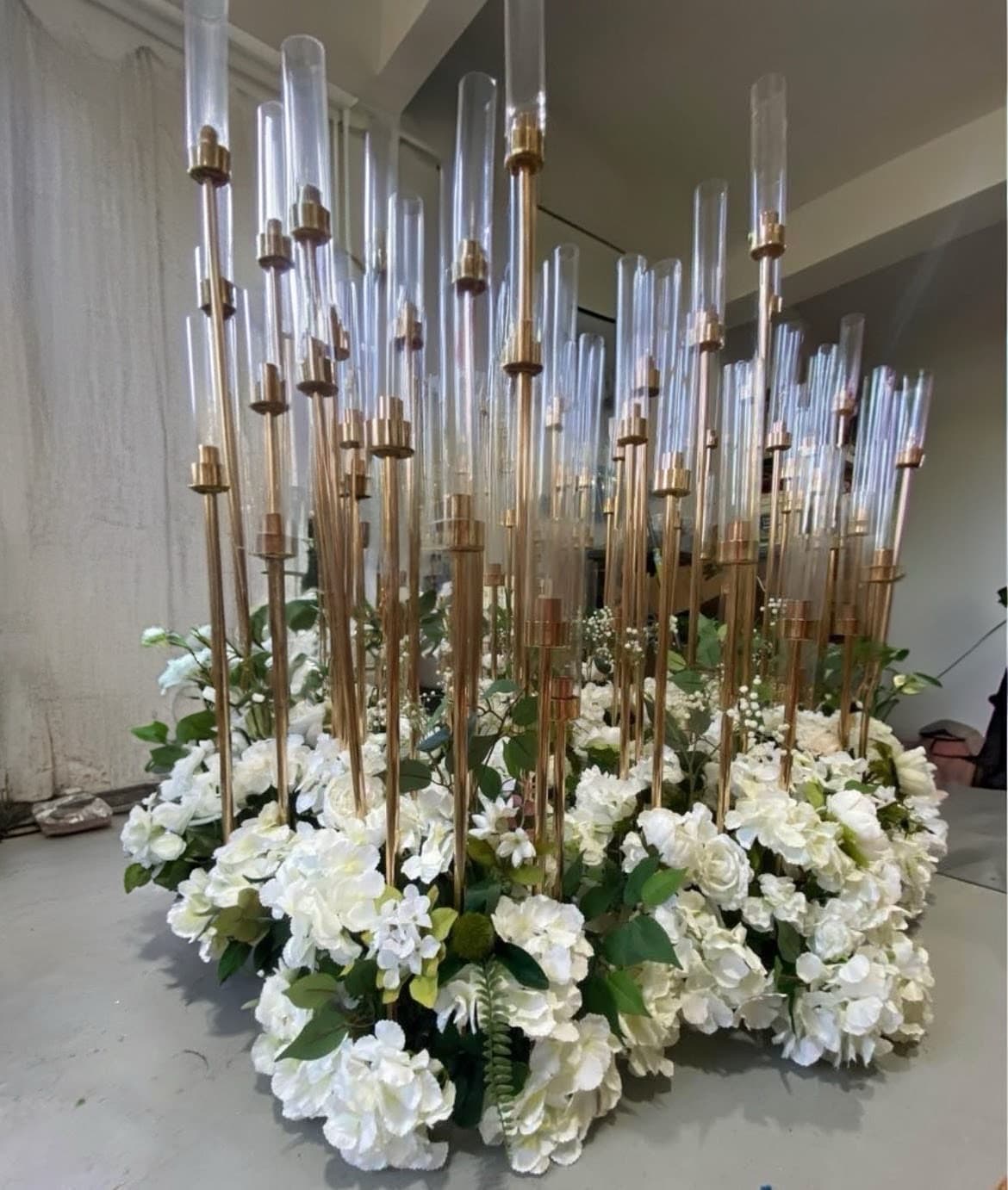 Large white floral arrangement featuring numerous tall gold candle holders with clear glass cylinders.