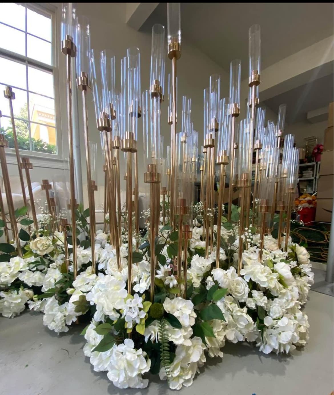 Tall gold candle holders with glass cylinders rising from a lush base of white flowers.