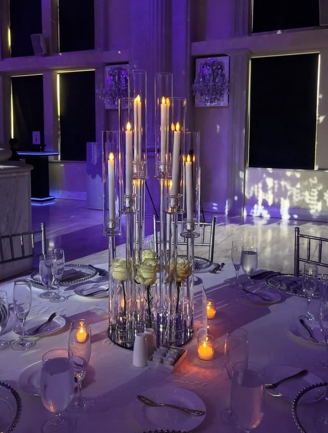 Tall glass candelabra with lit candles and white roses on a table with purple lighting.