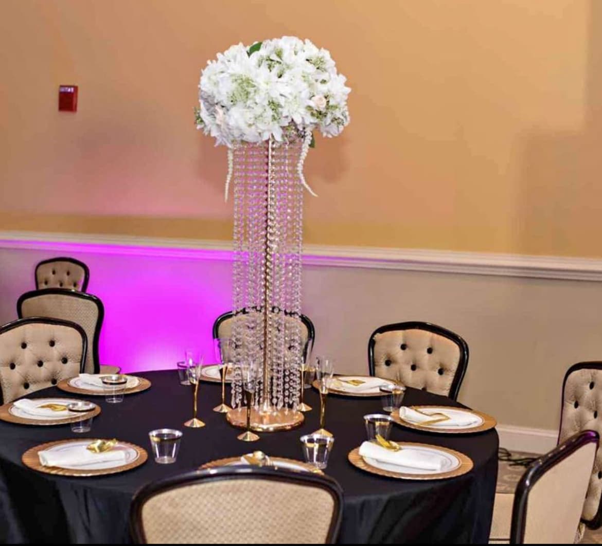 Elegant round table with black linens, gold chargers, and a tall crystal-draped white floral centerpiece.